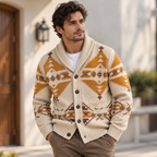A man wearing a cream-colored knit cardigan with a brown and ochre geometric pattern, styled over a white t-shirt and khaki pants, standing outdoors in front of a neutral wall.
